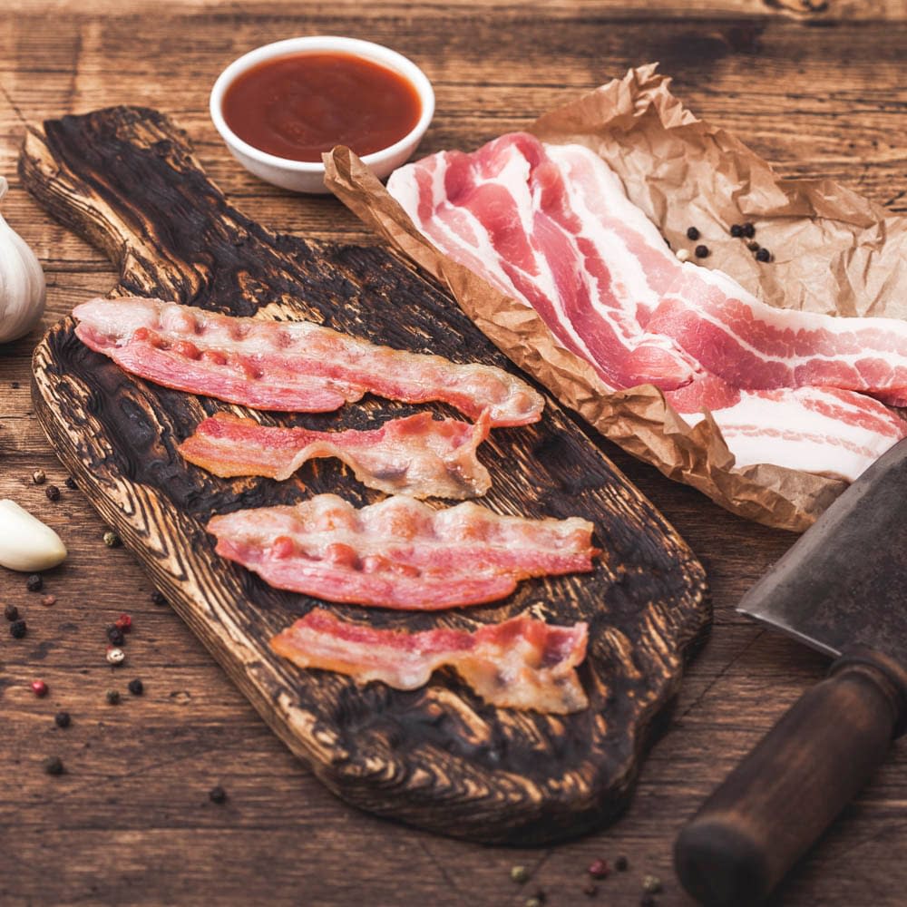 Grilled bacon strips on vintage wooden board with raw fresh smoked pork bacon on butchers paper with meat hatchet on wood board.